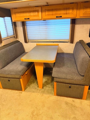 A compact RV interior with a central table, two cushioned seating areas, wooden cabinets, and a window with blinds.