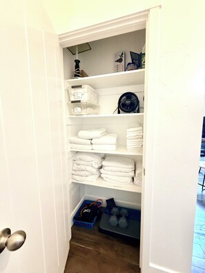 Een witte kast met planken, gevuld met opgevouwen handdoeken, een ventilator en opbergmanden.