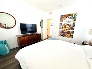 Ein Schlafzimmer mit einem Bett, einem Fernseher auf einer Holzkommode, einem großen abstrakten Gemälde und einem Spiegel an der Wand.