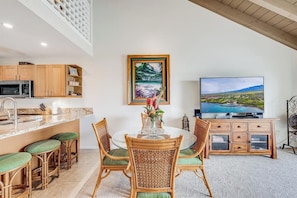 A modern kitchen with a microwave, wooden cabinets, and a dining area with wicker chairs and a glass table. - Experience Paradise, 2-Bed Kihei Condo with A/C and New June 2026 Ownership J214 (Kihei)