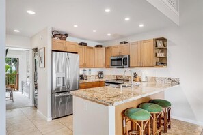 A modern kitchen with granite countertops, stainless steel appliances, wooden cabinets, and a central island with bar stools. - Experience Paradise, 2-Bed Kihei Condo with A/C and New June 2026 Ownership J214 (Kihei)
