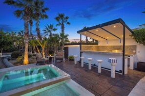 A modern outdoor kitchen area with a bar, seating, and a pool. - Luxury 4BR Villa in Pedregal | Minutes to Beach (Cabo San Lucas)