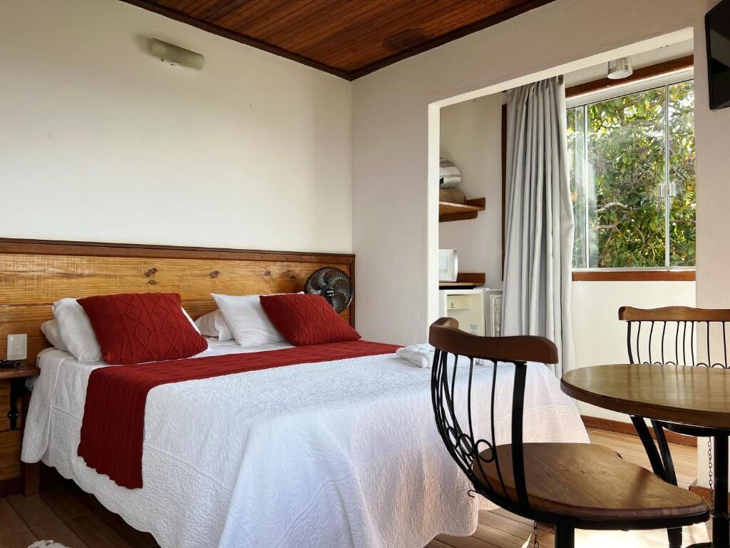 A bedroom with a wooden headboard, a bed with white bedding and red accents, a bedside table, a chair, and a window with curtains.