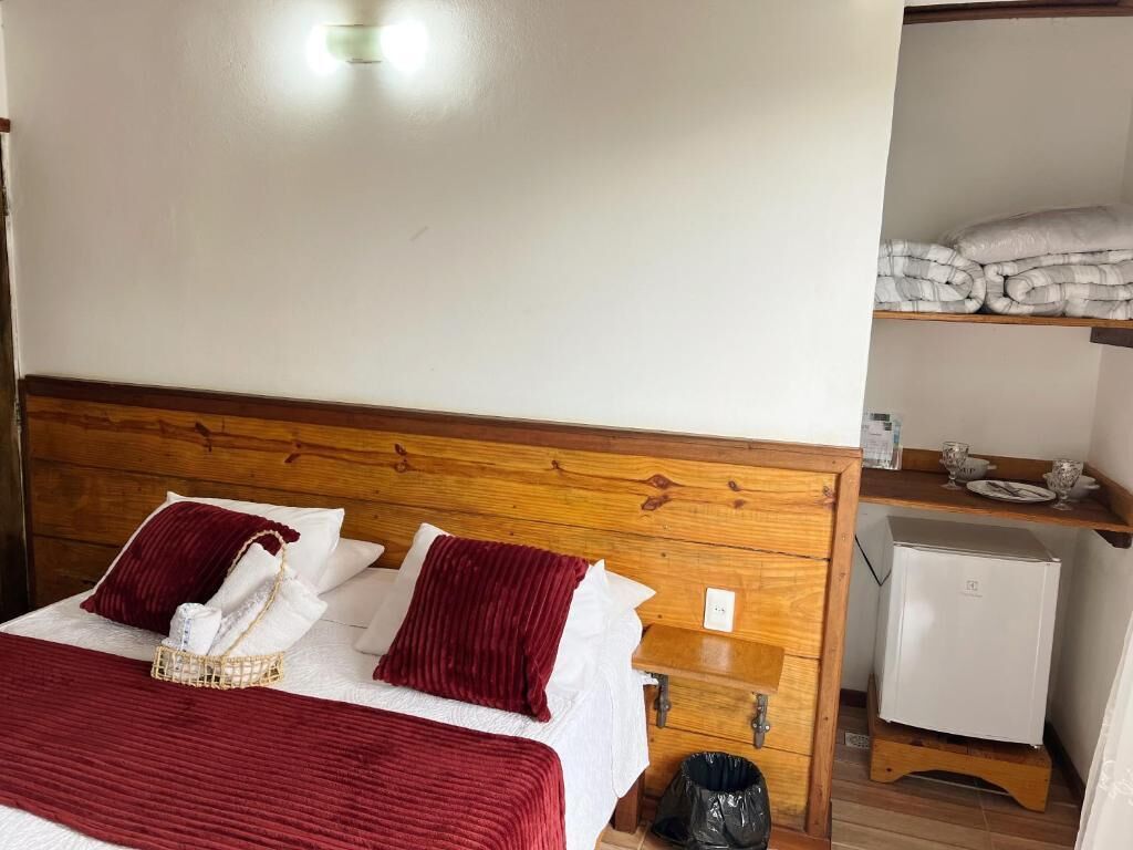 Habitación de hotel con cabecera de madera, cama con ropa blanca y almohadas rojas, mesita de noche con lámpara y refrigerador compacto.