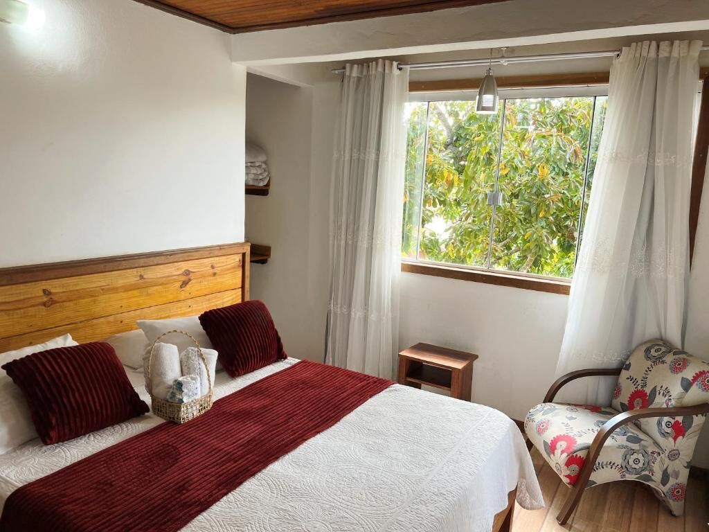 A bedroom with a wooden headboard, a bed with white and red bedding, a small bedside table, a chair with floral upholstery, and a window with curtains.
