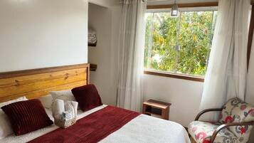A bedroom with a wooden headboard, a bed with white and red bedding, a small bedside table, a chair with floral upholstery, and a window with curtains.