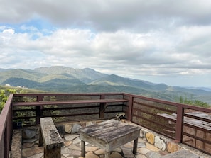 A wooden bench and table set on a stone patio with a view of green mountains.