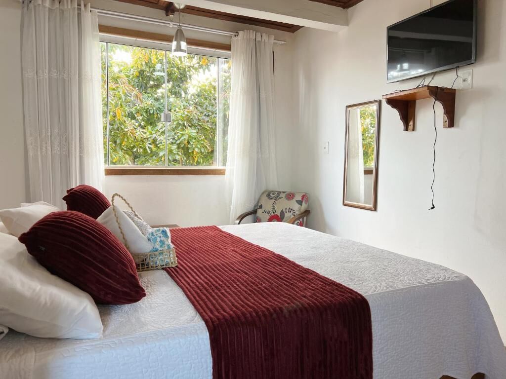 A bedroom with a bed, a window with curtains, a chair, a mirror, and a television mounted on the wall.
