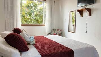 A bedroom with a bed, a window with curtains, a chair, a mirror, and a television mounted on the wall.