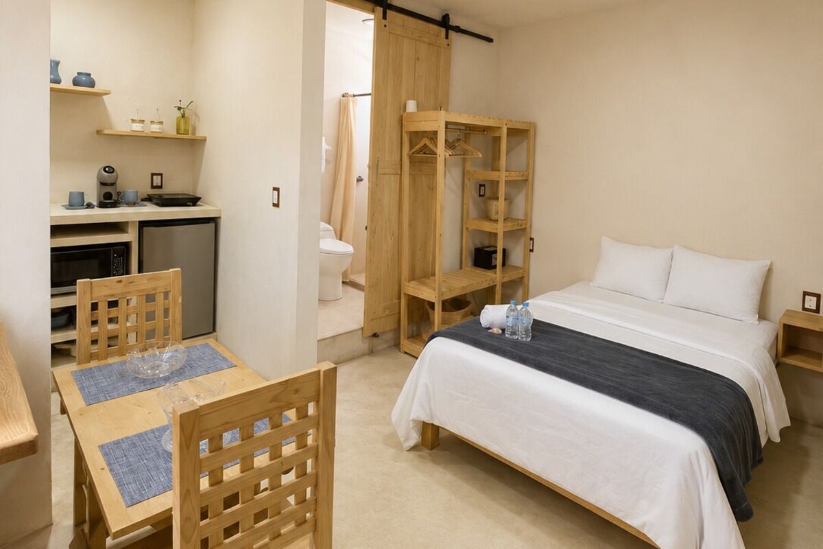 A compact hotel room with a bed, a small kitchenette, a wooden dining table with chairs, and a wardrobe.