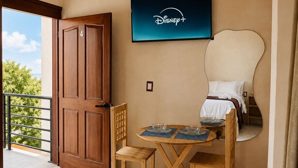 A hotel room with a balcony, a wooden door, a television mounted on the wall, a bed with a white headboard, and a small table with chairs. - Casa Maakja (Ixmiquilpan)