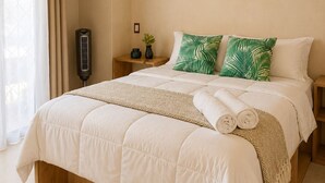A neatly made bed with white bedding, two green leaf-patterned pillows, a bedside table with a small plant, and a wall-mounted heater. - Casa Maakja (Ixmiquilpan)