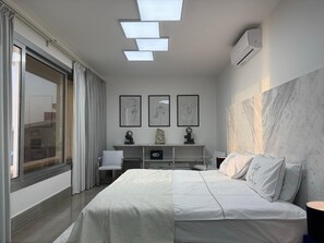 Ein modernes Hotelzimmer mit einem großen Bett, einem Schreibtisch und einem Sessel. An der Wand hängen drei gerahmte Kunstwerke, eine Marmorwand und ein großes Fenster mit Vorhängen.