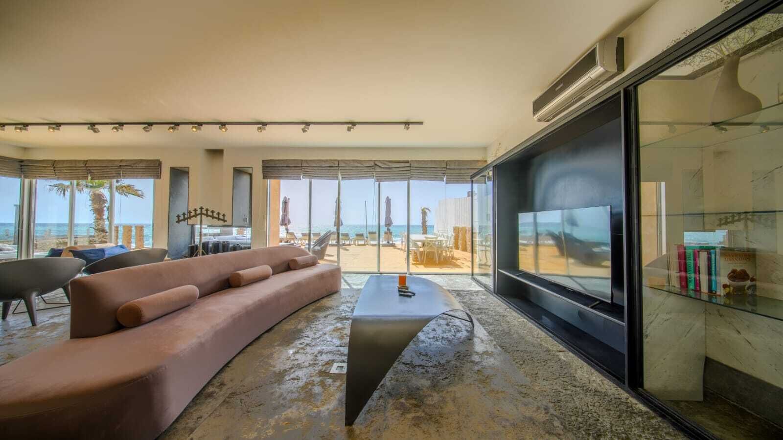 Ein modernes Wohnzimmer mit einer großen Couch, einem Couchtisch und einem Fernseher. Große Fenster bieten Blick auf den Strand.