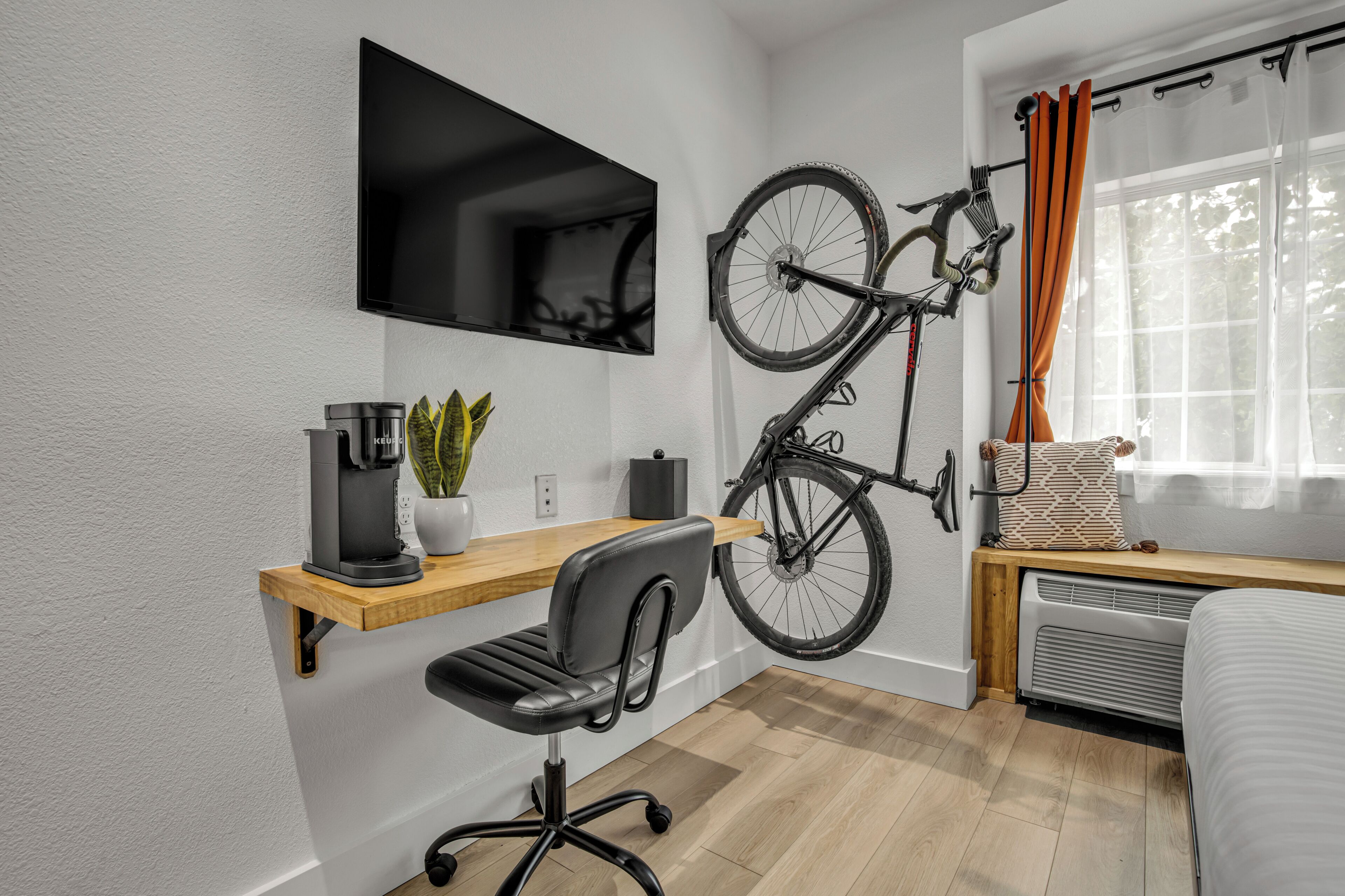 A modern room with a mounted TV, a bicycle, a desk with a chair, a potted plant, and a window with curtains.