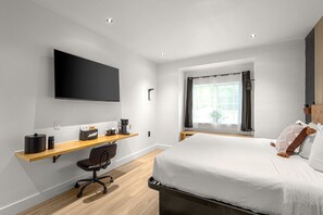 Ein modernes Hotelzimmer mit einem großen Bett, einem Schreibtisch mit Stuhl, einem an der Wand befestigten Flachbildfernseher, einem Fenster mit Vorhängen und einem kleinen Tisch mit einer Kaffeemaschine.