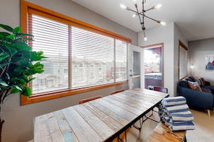 Un comedor con una mesa y sillas de madera, ventanas grandes con persianas, una planta en maceta y un sofá azul al fondo.