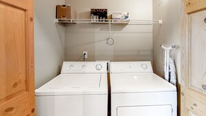 Uma área de lavanderia com duas máquinas de lavar brancas, uma prateleira com uma caixa Revlon e uma caixa Black & Decker, e uma porta de madeira.