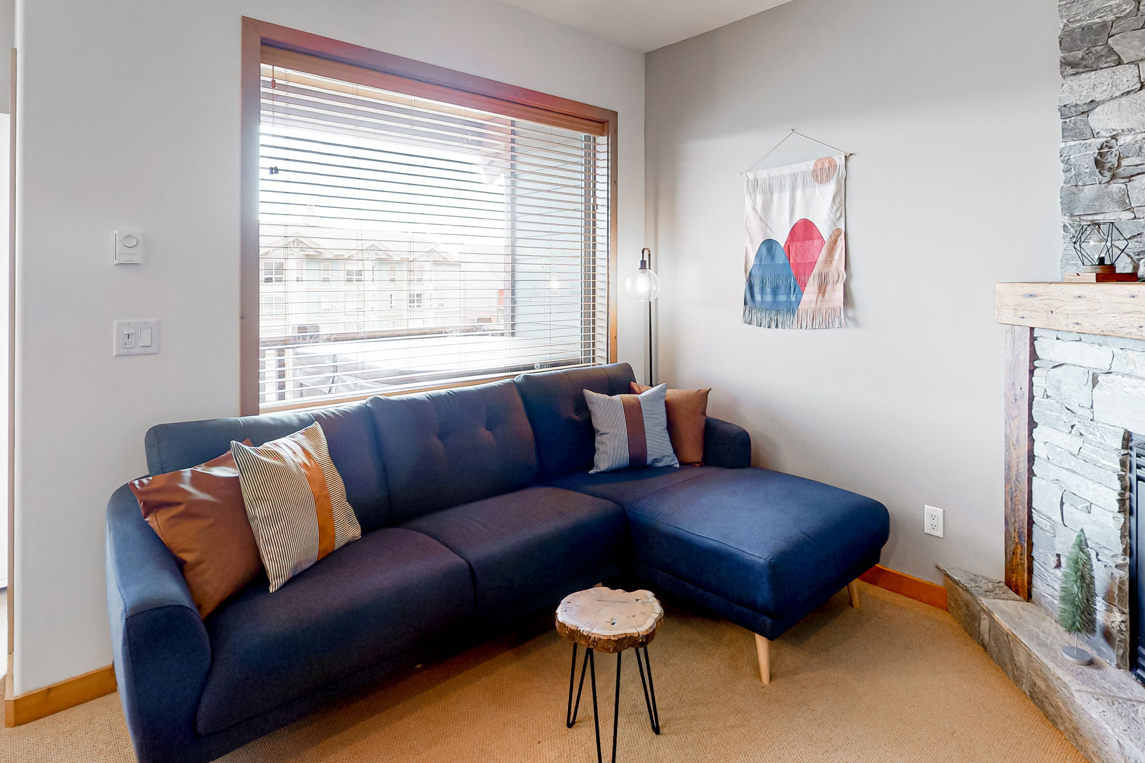 Ein modernes Wohnzimmer mit einem dunkelblauen Ecksofa, einem kleinen runden Holzschemel und einem Kamin aus Stein.