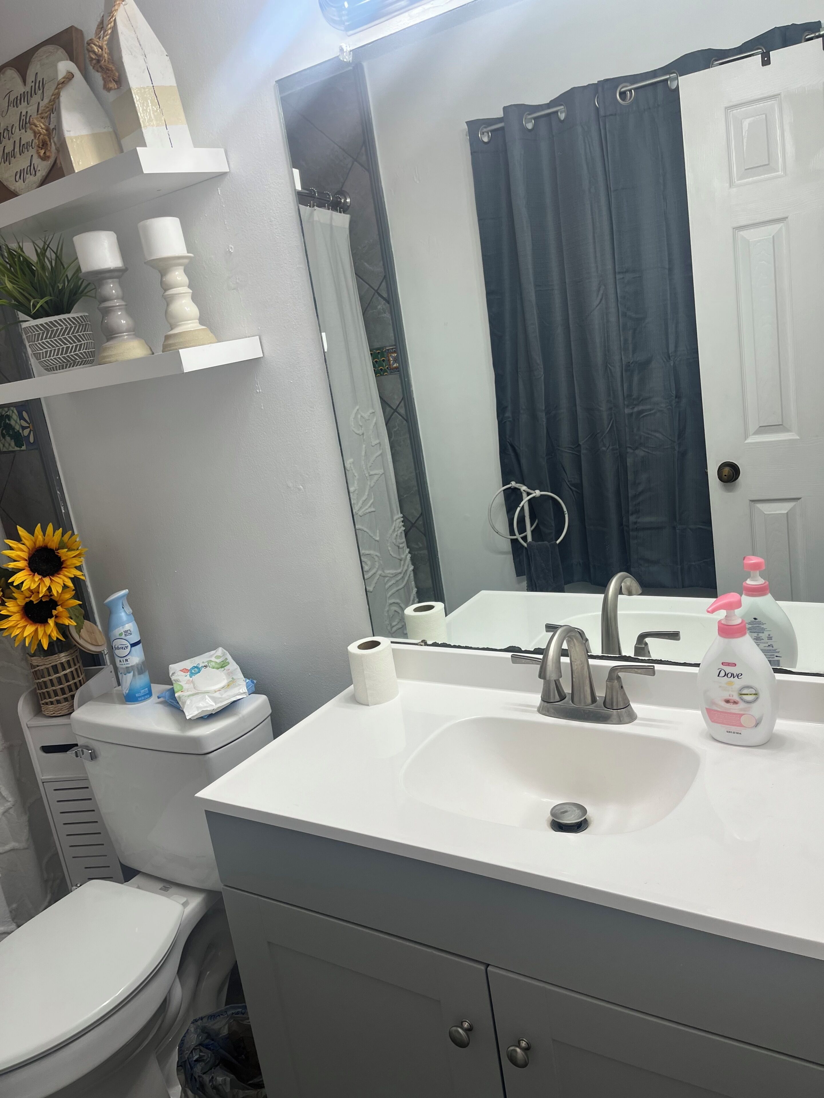 A bathroom with a white sink, a mirror, a dark curtain, a toilet, and a shelf with decorative items.