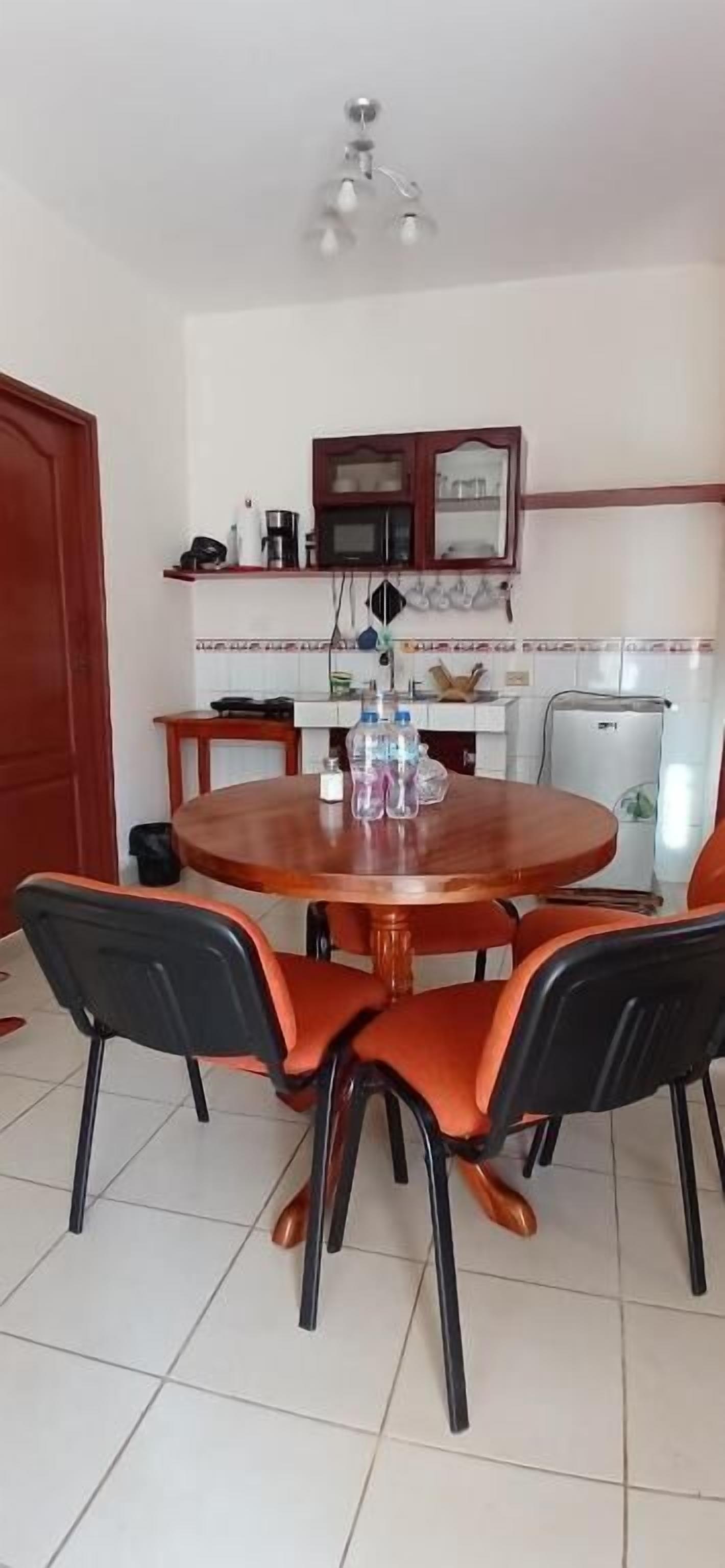 Un área de comedor con una mesa redonda de madera, sillas negras y anaranjadas, una cocina al fondo y una lámpara en el techo.