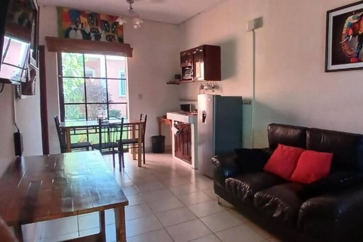A living room with a dining table, a sofa with a red pillow, a kitchen area with a refrigerator and cabinets, and a window with a view of greenery.