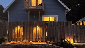 Una casa de dos pisos con balcón, cerca viva de madera y un jardín delantero bien iluminado.