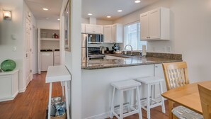 Una cocina moderna con armarios blancos, encimera de granito y una zona de comedor con mesa de madera y taburetes blancos.