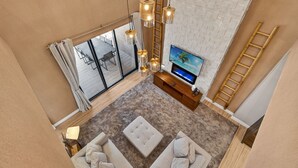 Ein modernes Wohnzimmer mit einem großen Fernseher, beigen Sofas, einer Holz-TV-Bank und Blick ins Freie durch große Fenster.