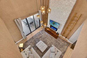 A modern living room with a large TV, beige sofas, a wooden TV stand, and a view of the outdoors through large windows. - Spacious 3BR/3.5BA Home Near Disney in Kissimmee (Kissimmee)