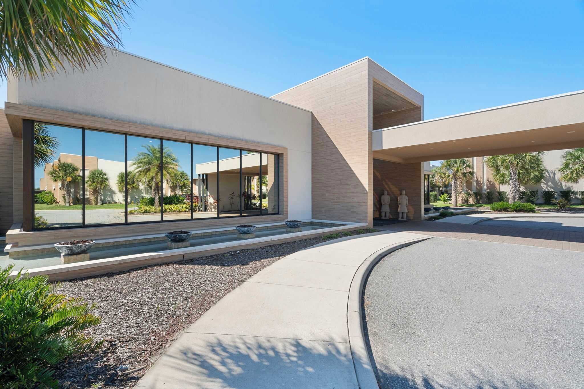 A modern building with large windows, a curved driveway, and a landscaped area with palm trees.