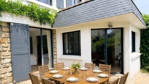 Ein runder Holztisch, gedeckt für sechs Personen, mit weißen Tellern und Gläsern, umgeben von Holzstühlen, in einer Außenpatio-Terrasse mit einer Steinsäule und einem Fenster mit Blick auf einen Garten.
