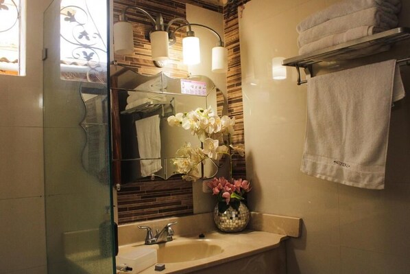 A bathroom with a glass shower enclosure, a sink with a mirror, and a vase with flowers. - Hostal Casa Las Flores (Masaya)