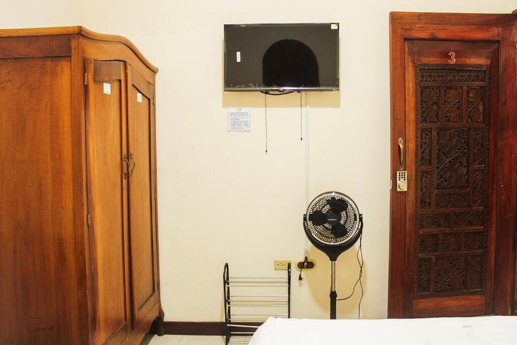 A room with a wooden wardrobe, a wall-mounted air conditioning unit, a standing fan, and a wooden door with a number.