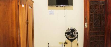 A room with a wooden wardrobe, a wall-mounted air conditioning unit, a standing fan, and a wooden door with a number.