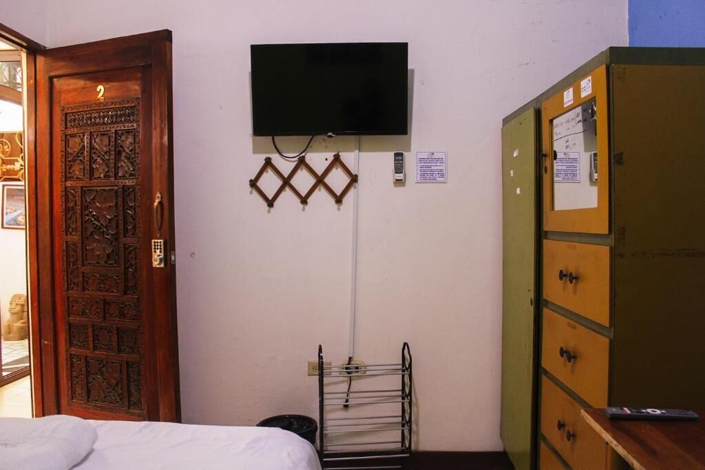 Una habitación con puerta de madera, televisor montado en la pared, un estante metálico y un archivador verde.