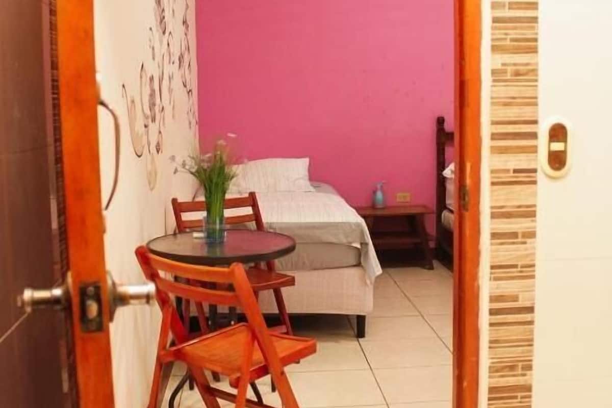 Una habitación con una cama, una mesa con sillas, una pared rosa y una puerta.