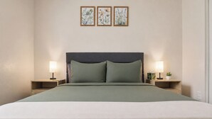 Una cama con cabecera oscura, dos mesitas de noche con lámparas, y tres láminas botánicas enmarcadas en la pared.