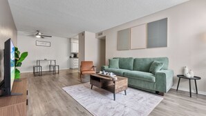 Una sala de estar con un sofá verde, una mesa de centro de madera, un mueble para televisión, un ventilador de techo y una cocina al fondo.
