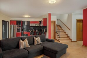 A living room with a sectional sofa, a kitchen in the background, and a staircase leading to another floor. - Casa Santo António –  House with Pico Views (Campo de Cima)