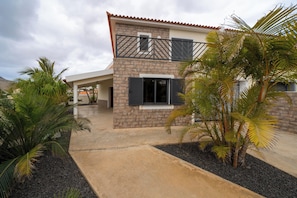 Uma casa de dois andares com fachada de pedra, uma varanda com grades de metal e uma palmeira em primeiro plano.