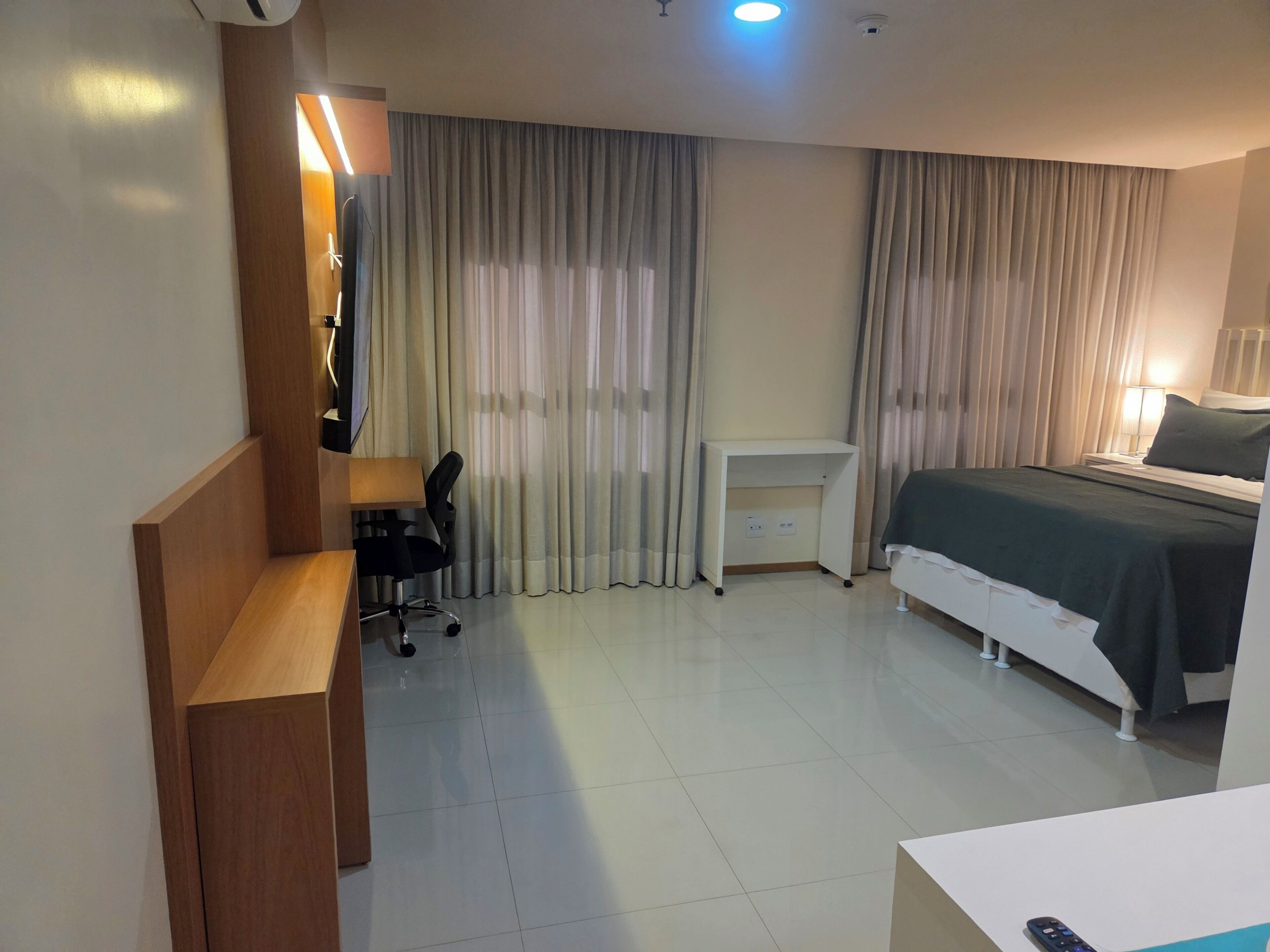 Quarto de hotel com cama, escrivaninha, cadeira e televisão.