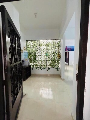 Un pasillo con piso de baldosas, una pared decorativa con follaje y una reja de metal negro a la izquierda.