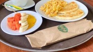 Una bandeja con huevos revueltos, una taza de café, rodajas de plátano y una bolsa de papel sobre una mesa de madera.