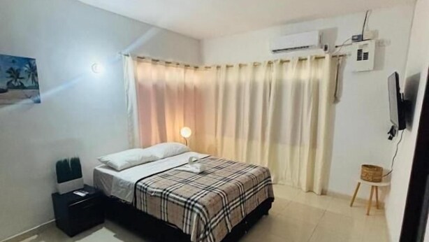 Un dormitorio con cama, mesita de noche, silla, televisor y ventana con cortinas.