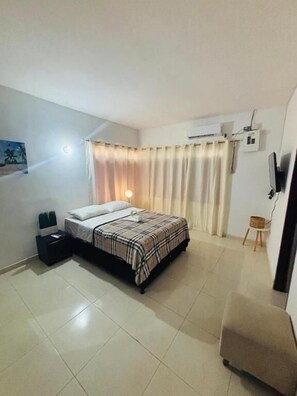 A bedroom with a bed, a nightstand, a chair, a TV, and a window with curtains.
