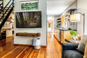 A modern living room with a large flat-screen TV, wooden flooring, a staircase, and a kitchen area with a lamp.