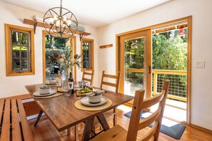 Un espace repas comprenant une table en bois, des chaises, un lustre et une vue sur la verdure grĂące Ă de grandes fenĂȘtres.