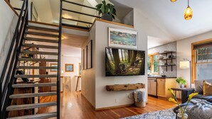 Un interior moderno con una escalera de madera, un televisor de pantalla plana montado en una pared blanca, una zona de cocina y un rincón de estar acogedor con un tapete estampado y un florero con flores.