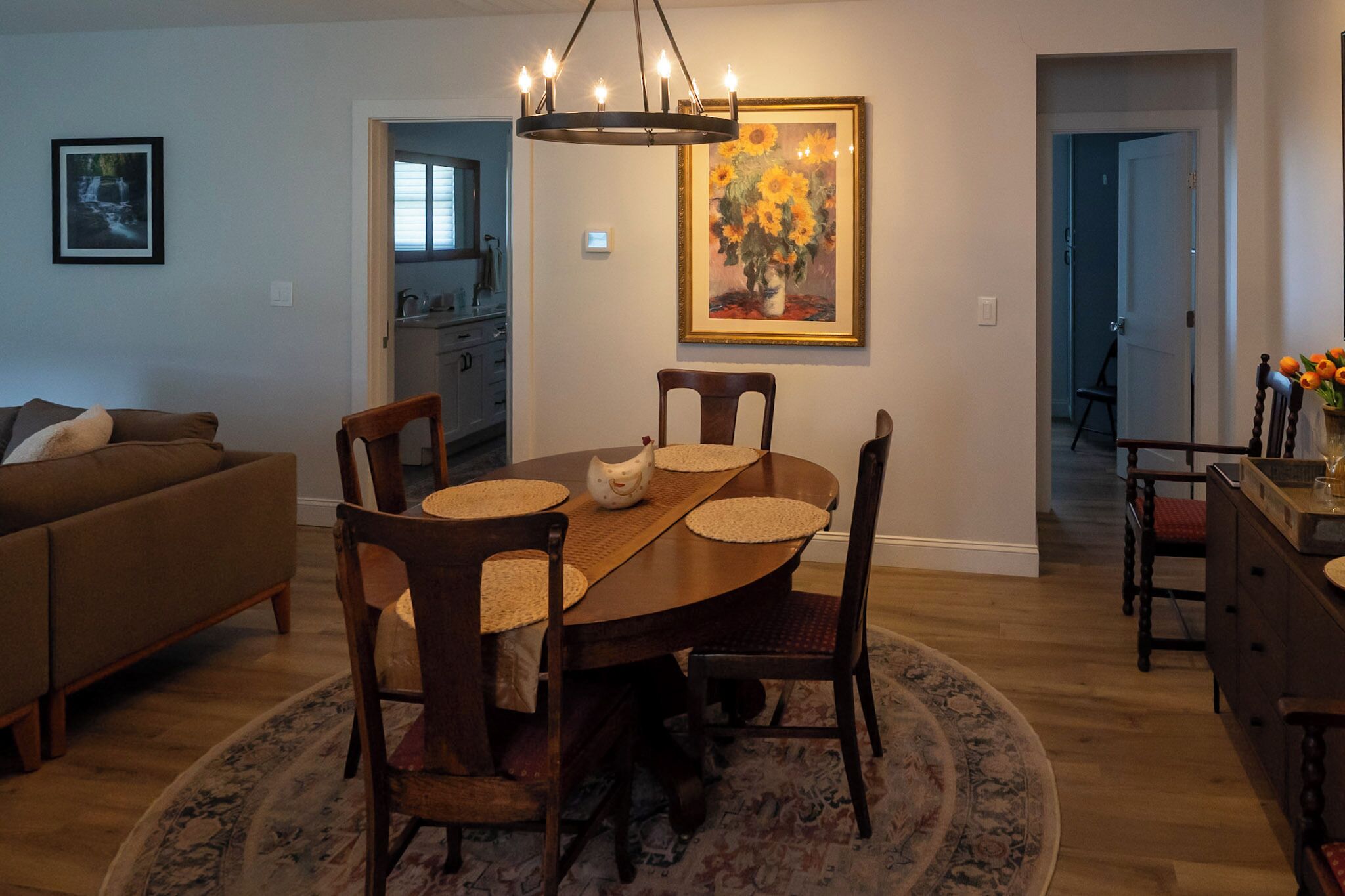 A dining area with a wooden table, chairs, and a chandelier. There is a painting on the wall and a kitchen in the background.